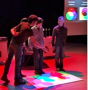 Illustration de la synthèse additive. Trois spots (vert, rouge et bleu) éclairent des élèves et projettent leurs ombres. L'élève du milieu, cachant le spot rouge, verra son ombre couleur cyan (addition du vert et du bleu).
